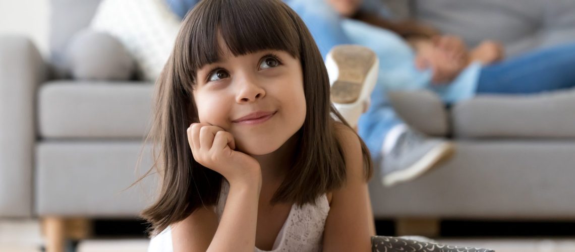 Close up little adorable thoughtful smiling daughter dreaming lying at cushion on warm floor with book in living room at modern home, resting married couple parents on background, focus on small kid